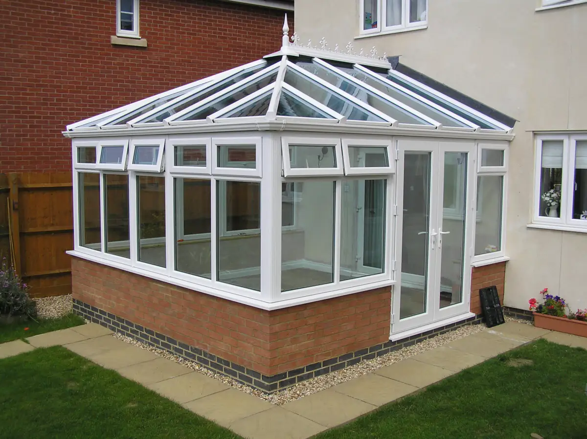 White uPVC Edwardian conservatory with a red brick dwarf wall and glass roof on the lawn behind an English home
