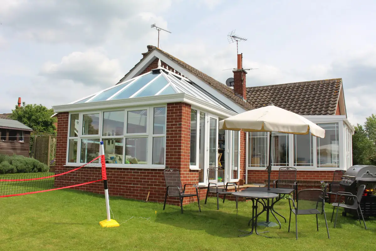 Large white uPVC conservatory with a hipped glass roof on a red brick base behind a bungalow, with garden furniture on the lawn