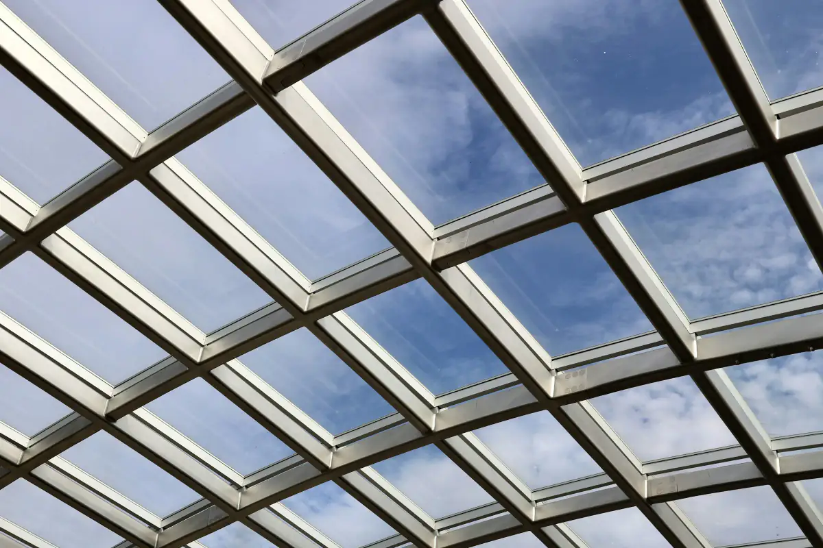 Looking up through a gridded glass roof at a bright blue sky with white clouds