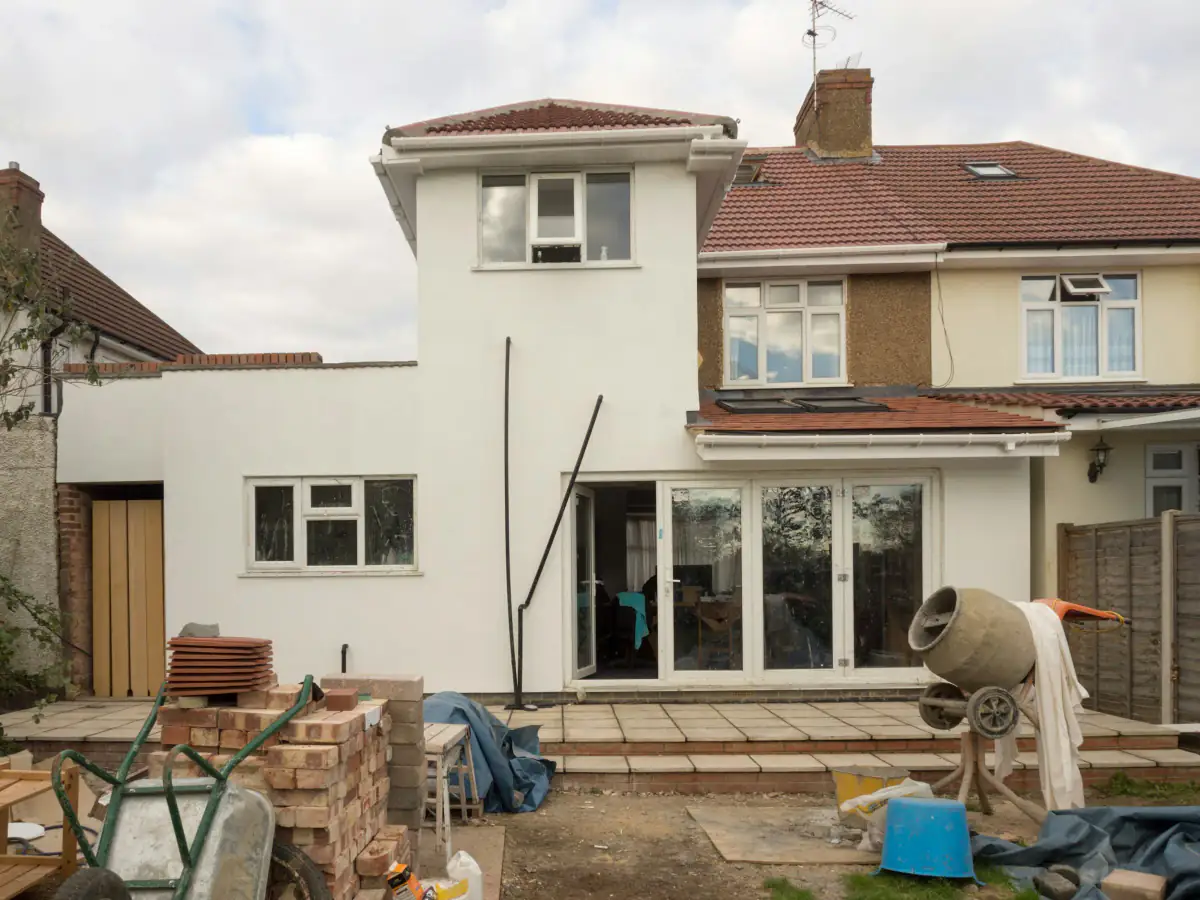 Rendered single storey rear extension under construction on a semi-detached house, with bricks, tiles and a cement mixer in the garden